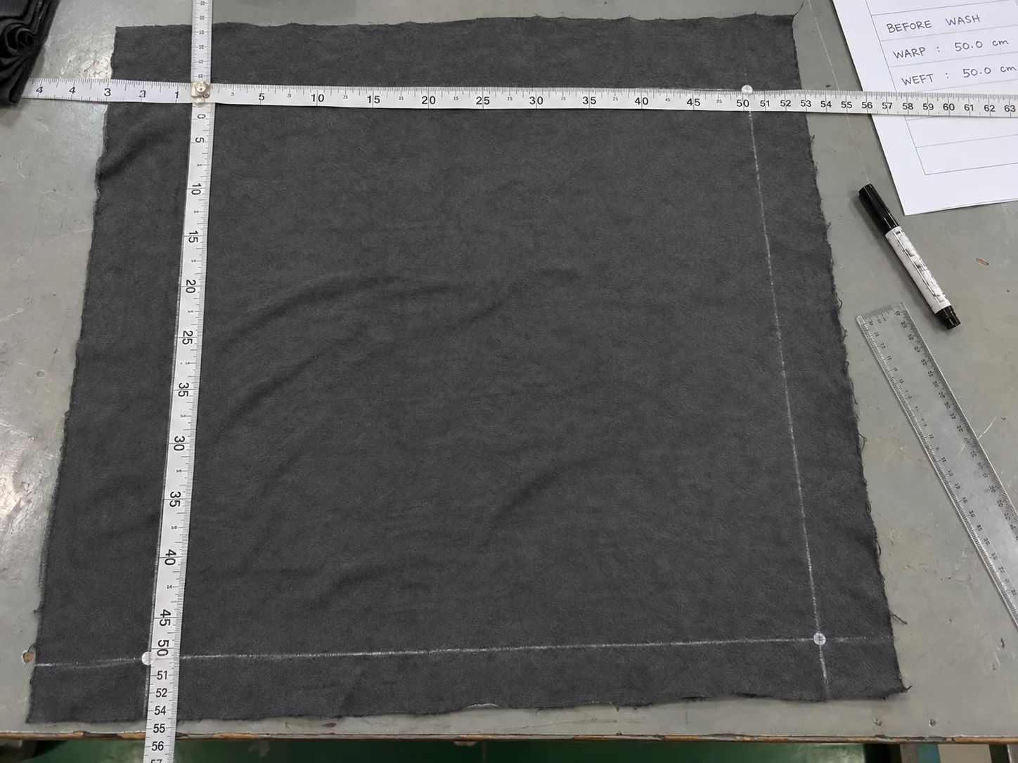 Dark knit fabric specimen marked with a square benchmark and measured in length and width before washing