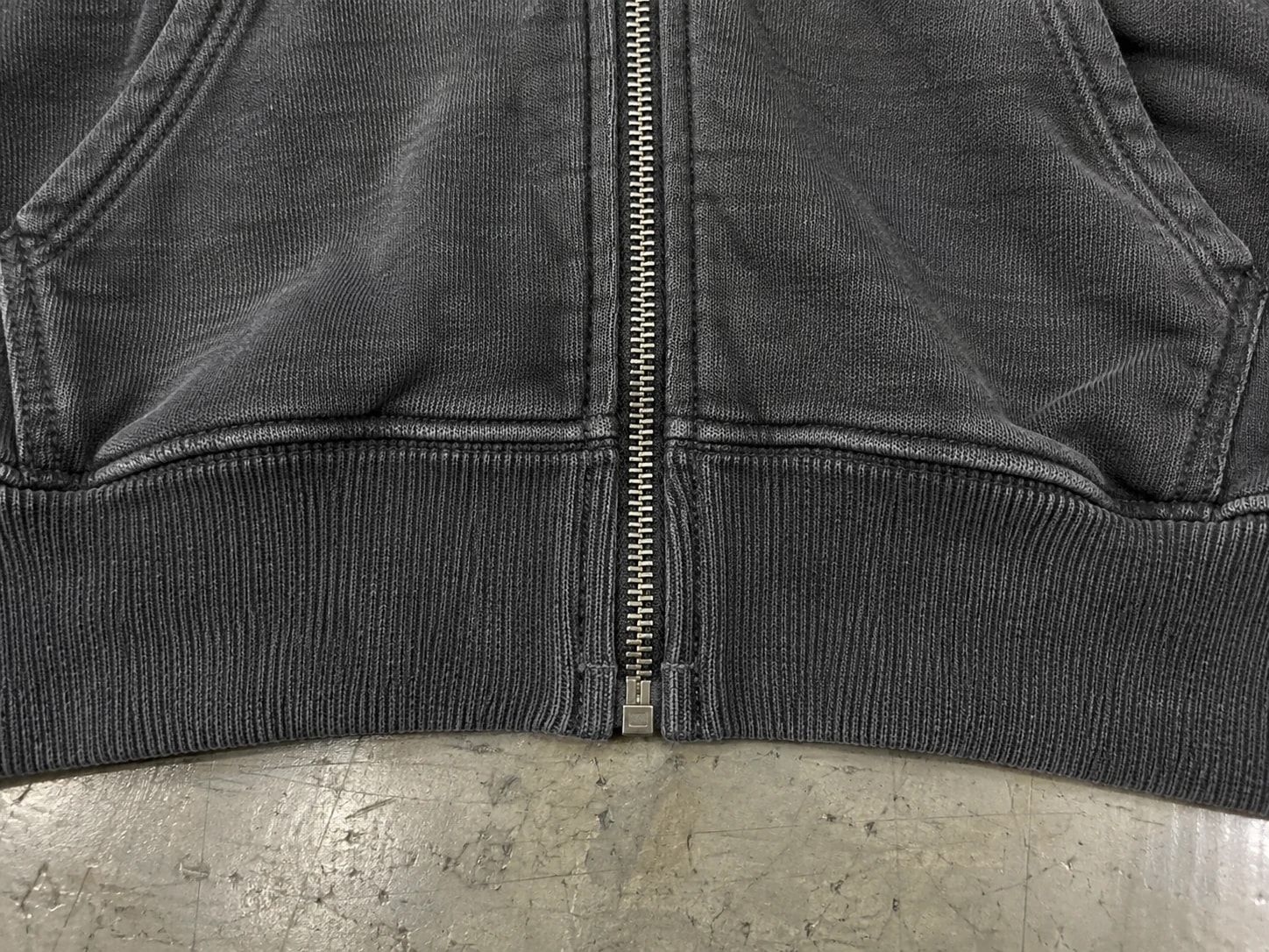 Metal zipper meeting the rib hem between split pockets on a washed dark hoodie