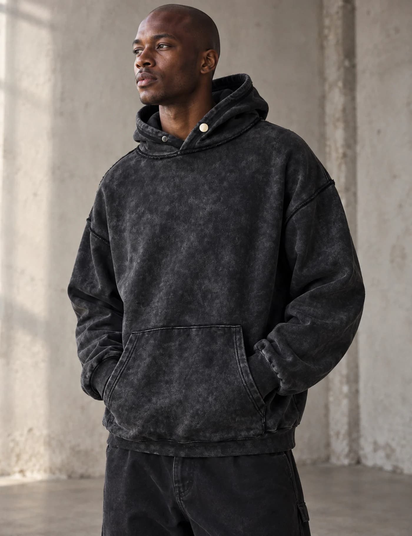 Male model wearing a black acid-wash pullover hoodie with a structured snap hood and small muted-red chest motif