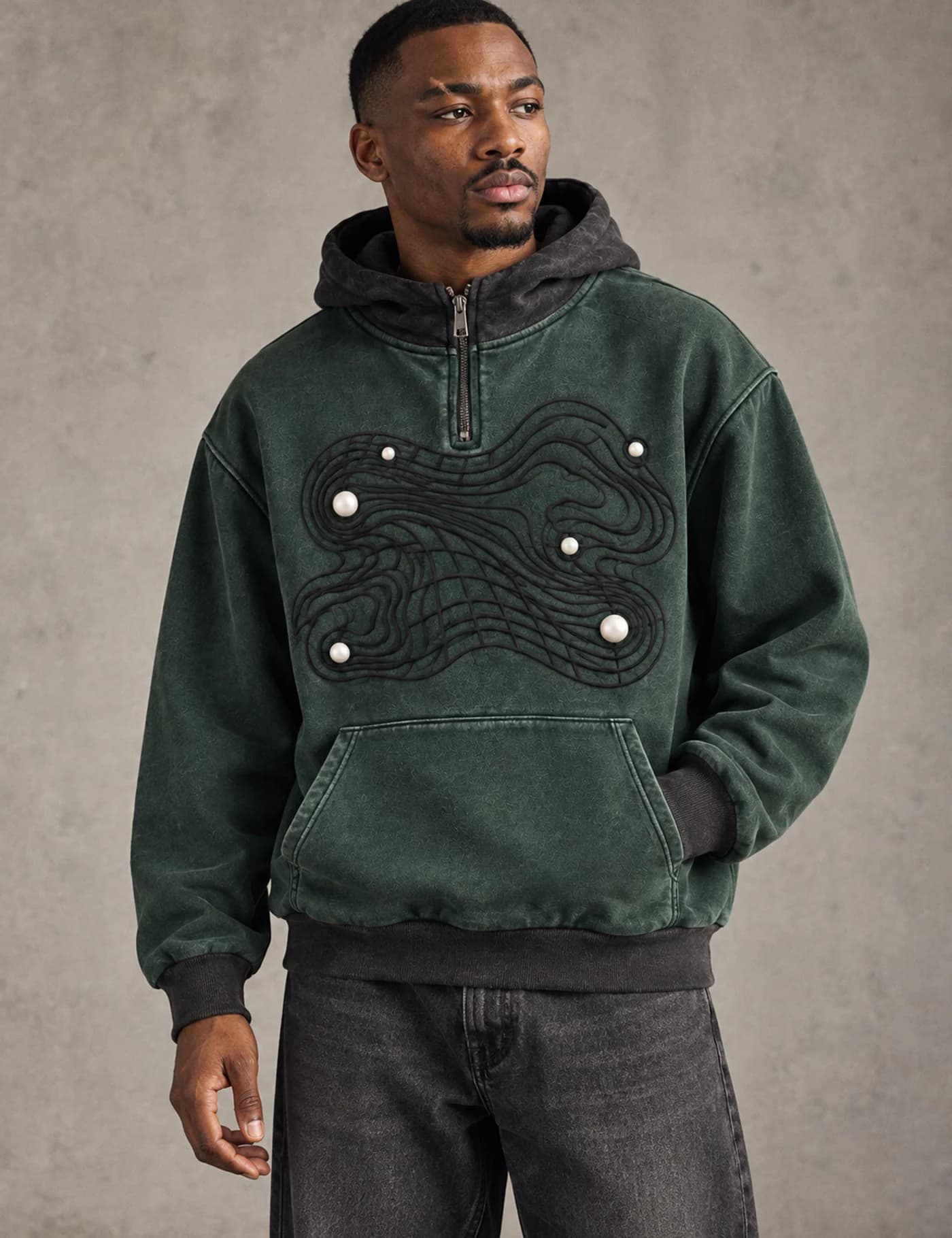 Male model wearing a washed green hooded quarter-zip sweatshirt with raised cord artwork and pearl-like accents