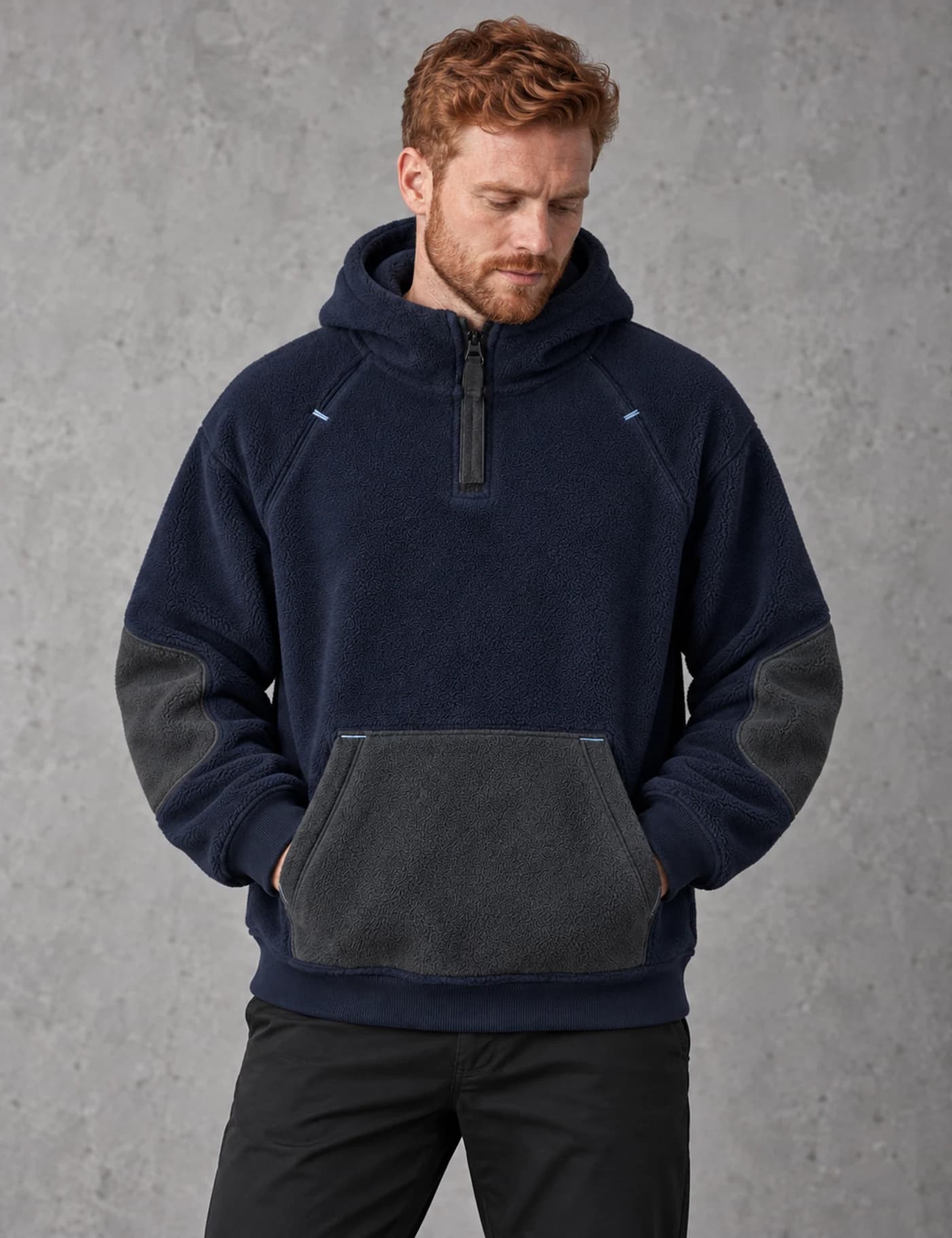 Red-haired European male model wearing a deep navy fleece half-zip hoodie with charcoal pocket and elbow panels