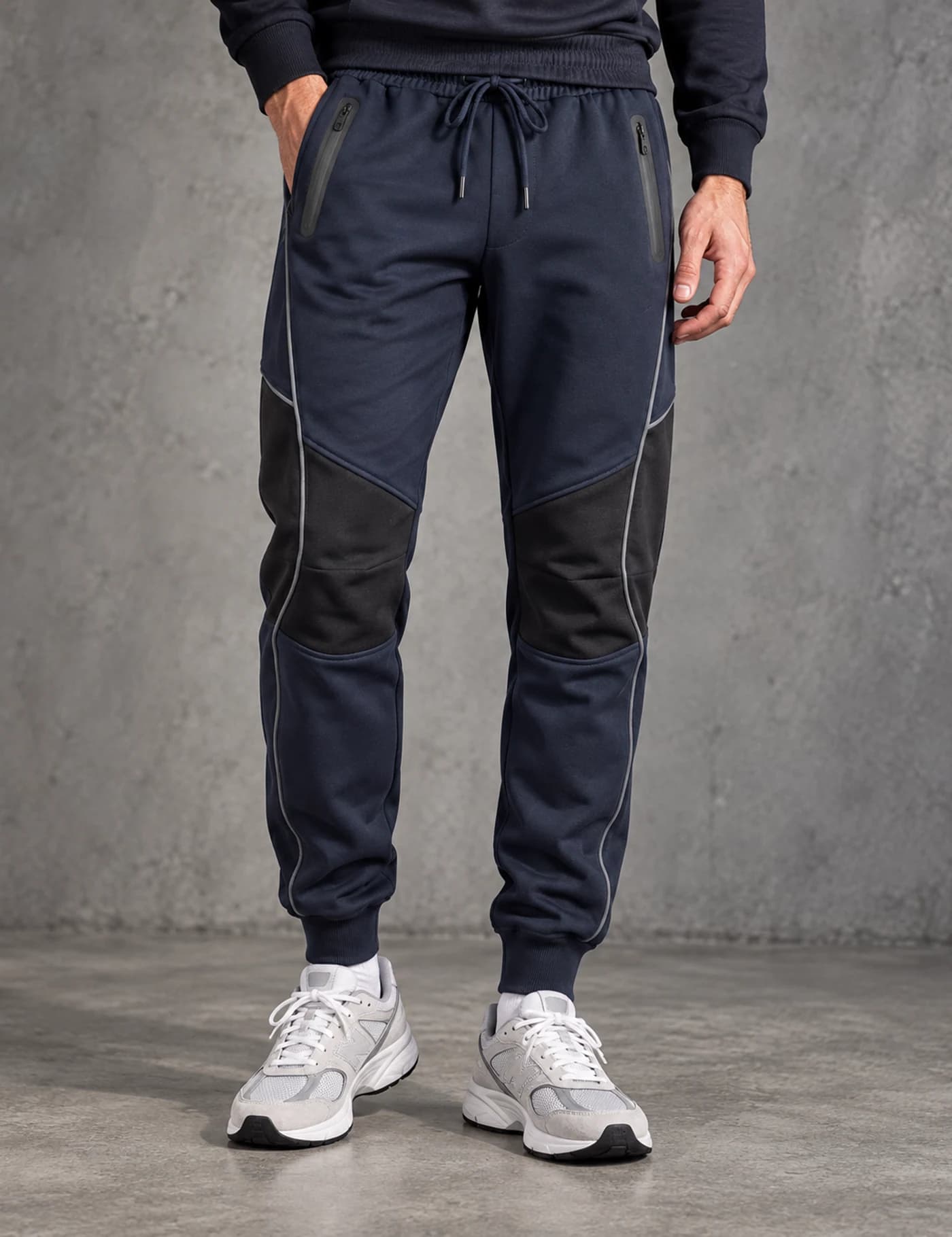 Male lower-body model wearing navy technical joggers with charcoal articulated panels and reflective piping