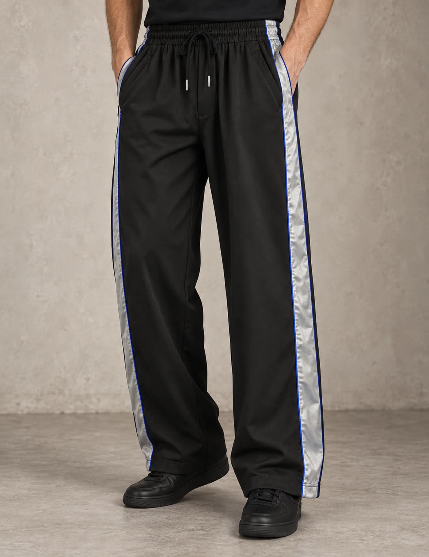Male lower-body model wearing black wide-leg track pants with broad silver reflective side stripes
