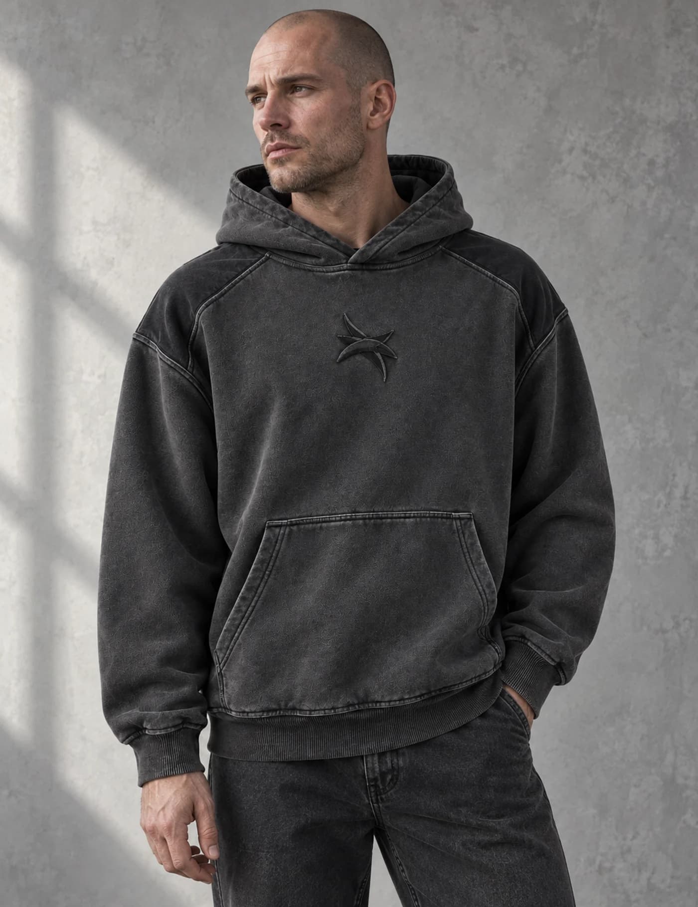 Shaved-head male model wearing a charcoal washed pullover hoodie with small tonal chest embroidery